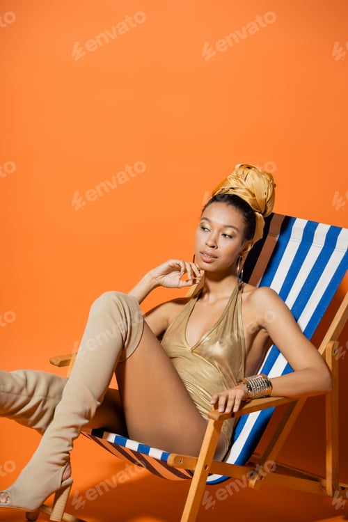 Preview: Trendy african american model in swimsuit and knee boots posing on deck chair on orange background