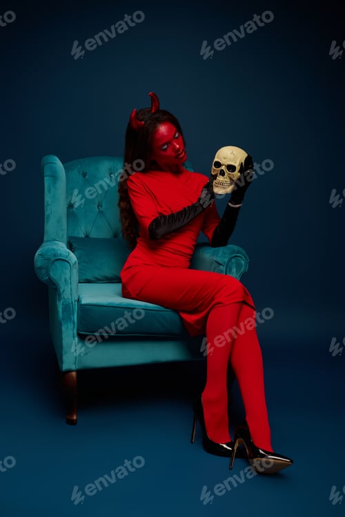 Preview: Visually captivating woman in a striking red Halloween costume holding a skull while seated