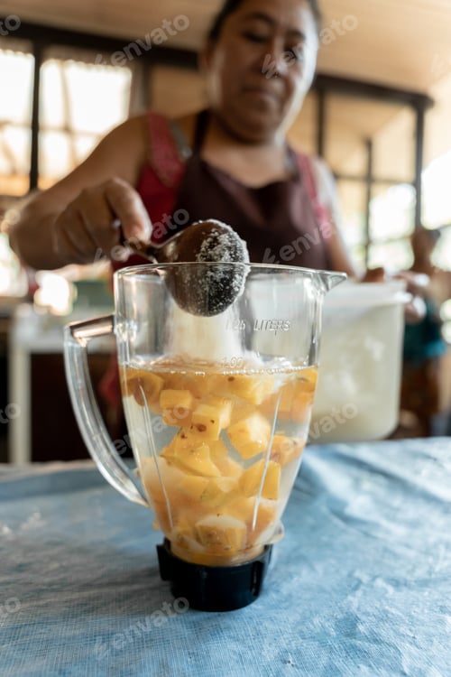 Preview: A young Hispanic woman is pouring sugar into a blender to make star fruit juice