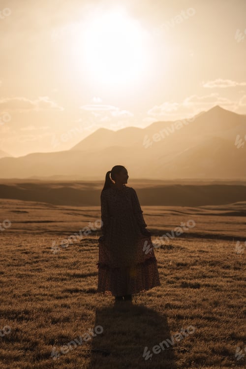 Preview: Asian woman in floral dress on golden field with snow mountain during golden sunrise at Sary Jaz