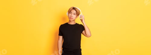 Preview: Young handsome asian guy with blond hair, take-off headphones to listen you, standing yellow
