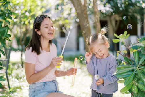 Preview: Laughing mother blowing bubbles at squinting smiling little girl in green garden