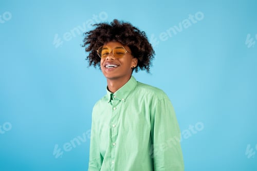 Preview: Portrait of happy african american teen guy in mint shirt and yellow sunglasses posing on blue