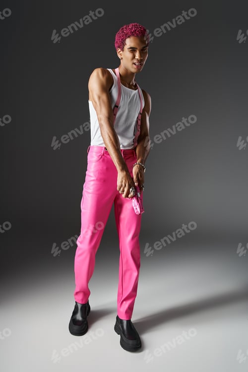 Preview: playful pink haired man in pants with suspenders posing with pink toy gun on gray background