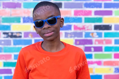 Preview: Wearing sunglasses, boy in orange shirt standing against colorful brick wall