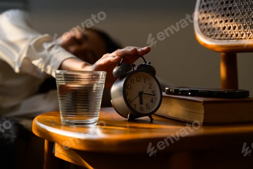 Preview: Woman stopping alarm clock in the morning near glass of water