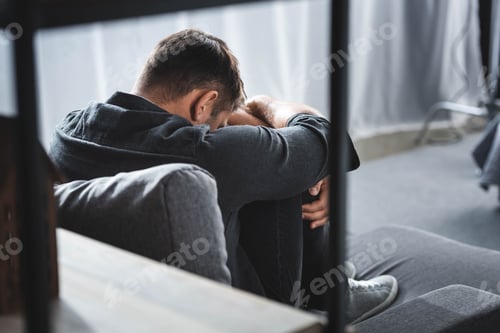 Preview: back view of man with panic attack crying and hugging legs in apartment