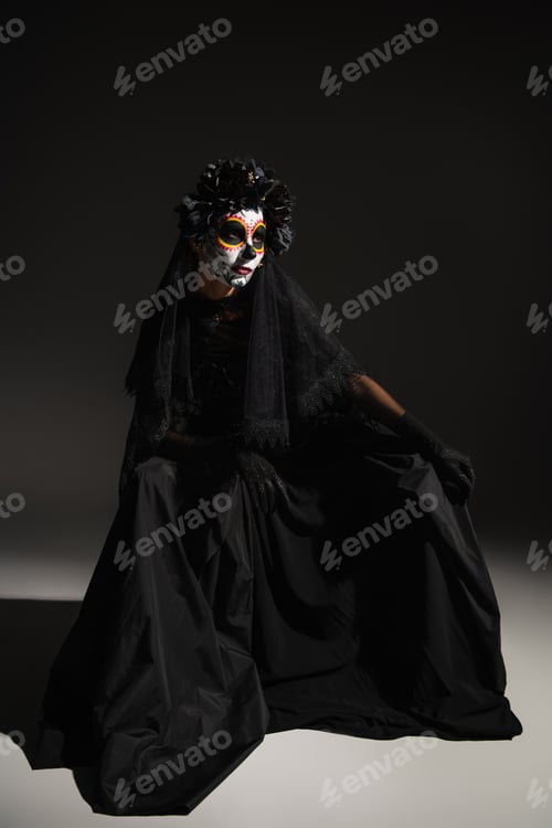 Preview: full length of woman in black witch dress and spooky halloween makeup sitting on dark background