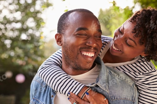 Preview: Smiling young black couple piggyback in garden, eyes closed
