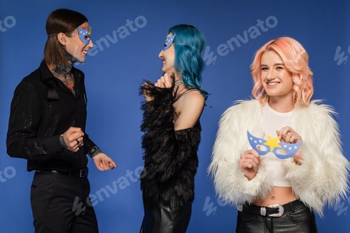 Preview: queer person holding party mask and looking at camera near nonbinary people dancing isolated on blue