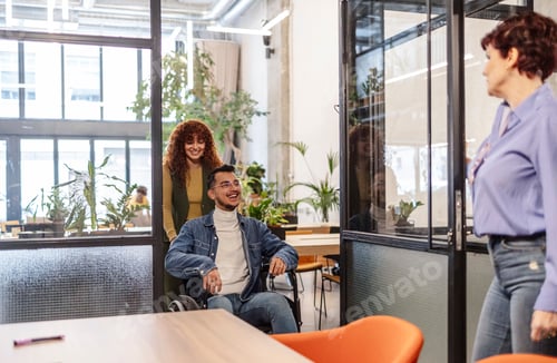 Preview: Businesswoman pushing disabled male coworker in wheelchair in modern office