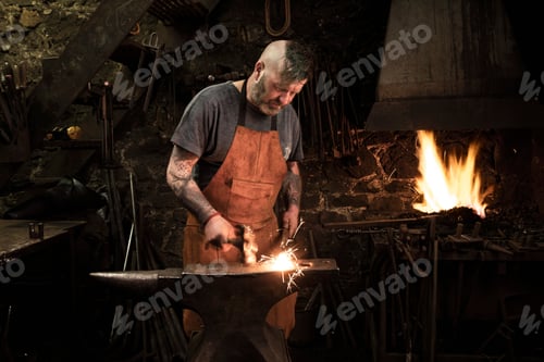 Preview: Mature male blacksmith hammering metal on anvil in workshop
