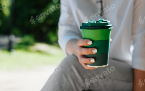 Preview: Unrecognizable woman holding disposable cup of coffee while sitting in park outdoors, close-up.