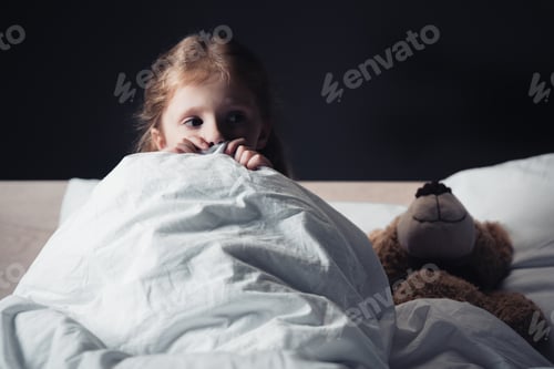 Preview: scared kid looking away while sitting under blanket near teddy bear isolated on black