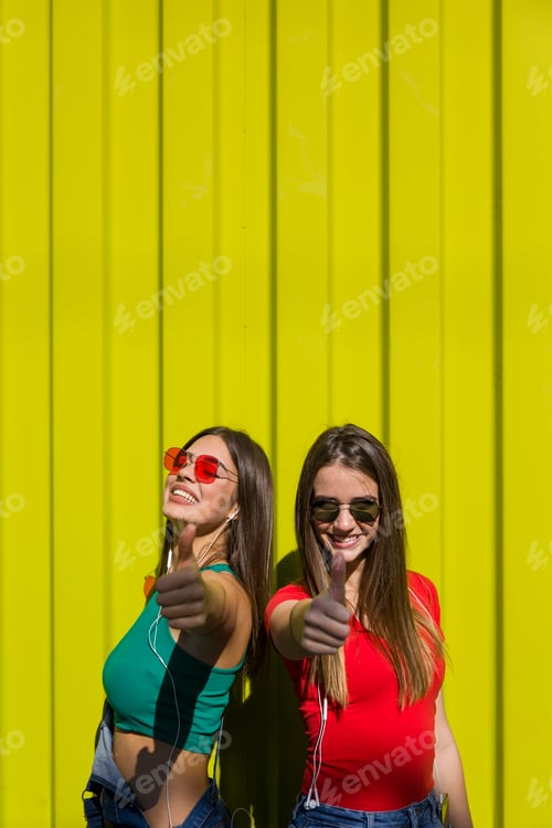 Preview: Portrait of two young happy women friends standing outdoor over yellow wall