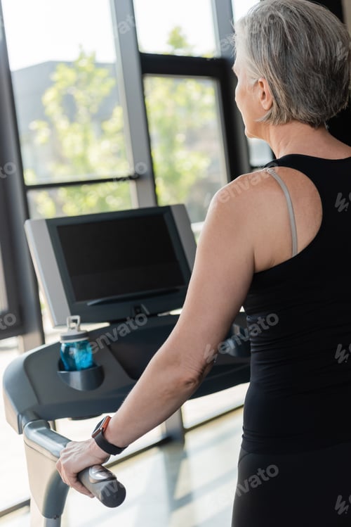 Preview: senior woman with grey hair running on treadmill next to sports bottle with water