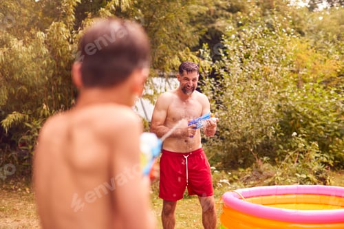 Preview: Father And Son Wearing Swimming Costumes Having Water Fight With Water Pistols In Summer Garden