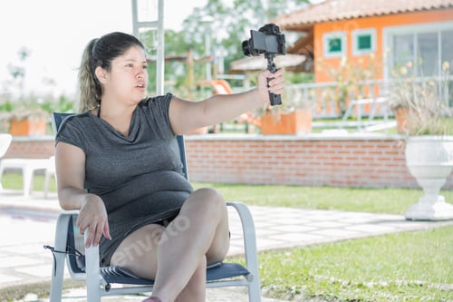 Preview: young latina woman blogger, sitting on a chair shooting a video of herself with camera in hand