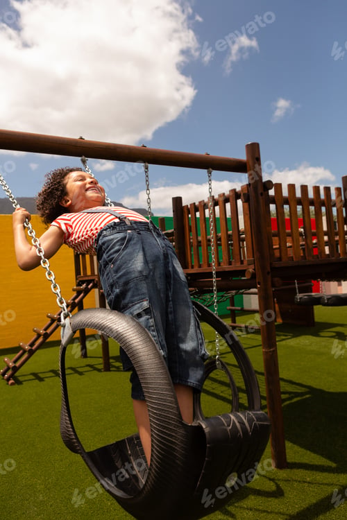 Preview: Side view of mixed-race schoolgirl playing on a swing at school playground on a sunny day