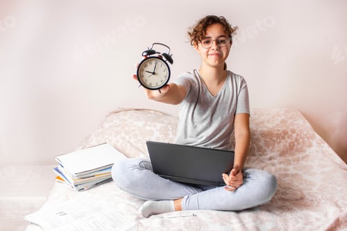 Preview: Happy funny teenager girl studying on the bed on computer and keeps alarm clock