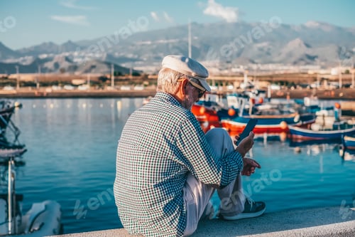 Preview: Handsome senior man in casual shirt and hat using phone while sitting at the sea port at sunset