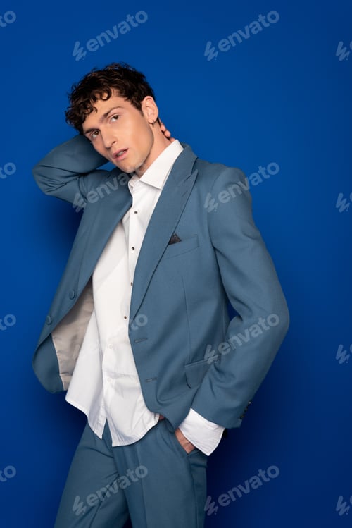 Preview: Fashionable curly model in suit and shirt touching neck on blue background