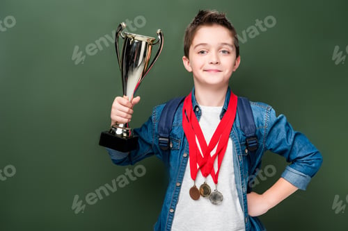Preview: schoolboy with medals holding winner cup near blackboard