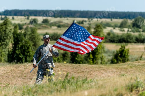 Preview: selective focus of handsome military man in uniform and cap holding american flag in summertime