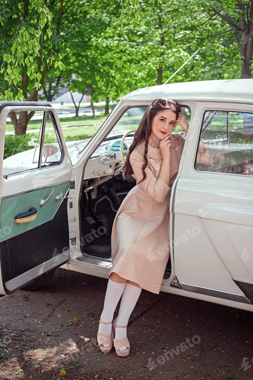 Preview: A pretty girl in a pink dress and with a retro hairstyle sits in a retro car with an open door