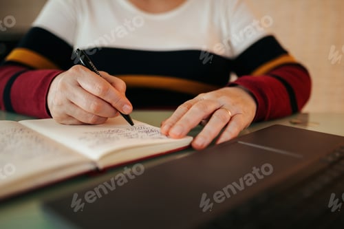 Preview: Person writing notes in a notebook while sitting at a desk with a laptop