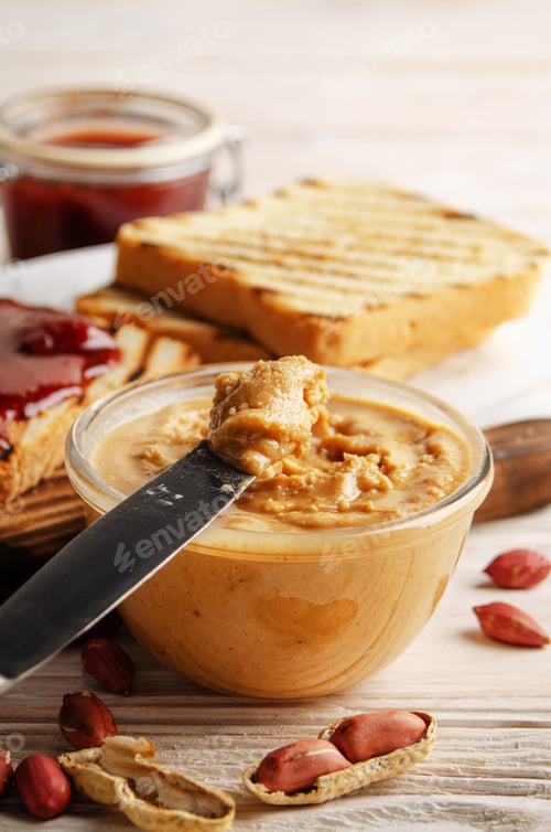 Preview: Low angle view at glass bowl with peanut butter on white wooden table with toasts and jam aside