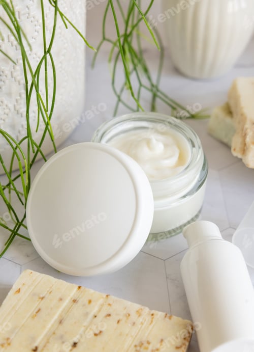 Preview: Opened cream jar with white blank lid on marble near green plant close up, cosmetic mockup
