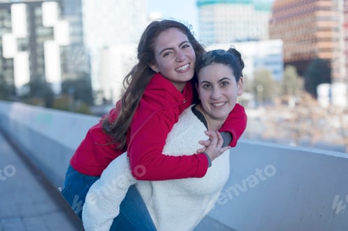 Preview: Front view of best smiling friends girls piggyback outdoors in the city