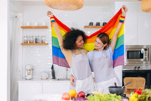 Preview: LGBT couple women happy celebrate pride month together with rainbow flag in kitchen cooking at home