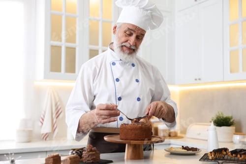 Preview: Elderly pastry chef with scoop and spoon of melted chocolate making cake at table in kitchen