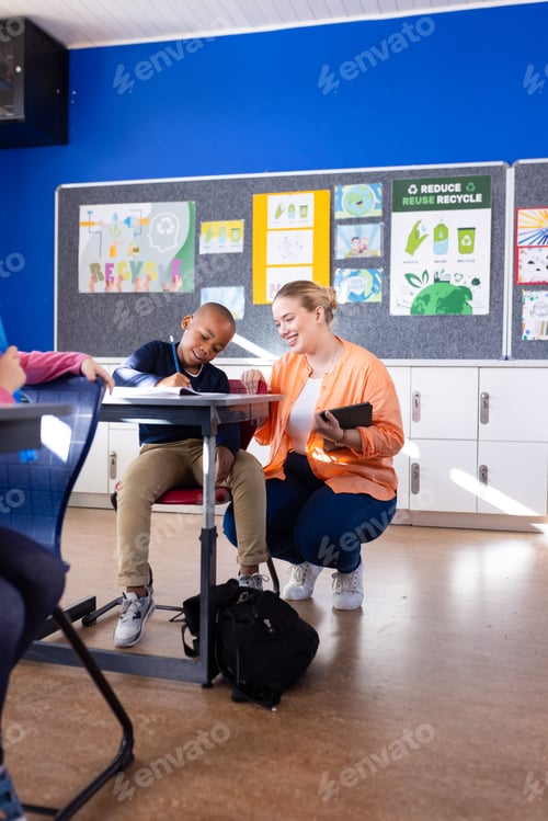 Preview: In school, female teacher helping african american boy with classwork at desk, smiling together