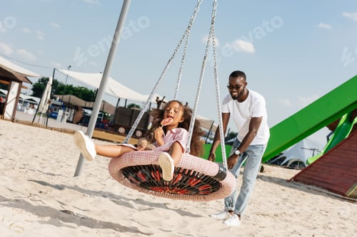 Preview: happy african american daughter with lollipop on spider web nest swing at amusement park