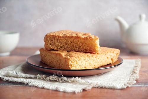 Preview: Two slices of sponge cake resting on a brown plate with teapot and cup in background