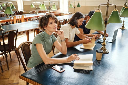 Preview: Portrait of beautiful girl with book dreamily studying with students in library of university