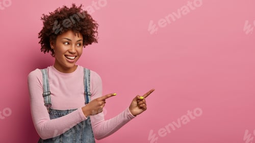 Preview: Studio shot of delighted dark skinned woman indicates at upper right corner, advertises new product