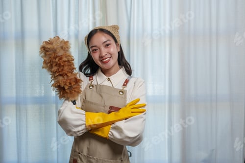 Preview: Happy housekeeper wearing yellow gloves and an apron, holding a feather duster and smiling with cros