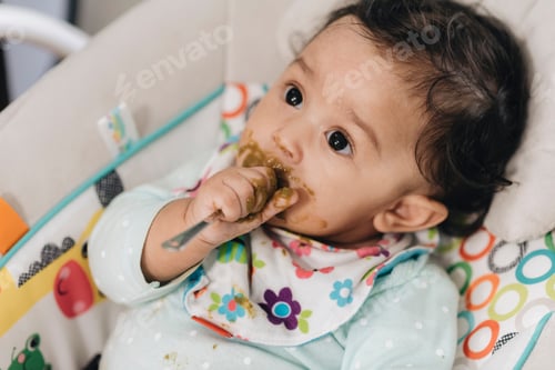Preview: Diverse toddler girl at home eating jar food with messy face