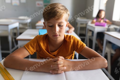 Visualização: Estudante caucasiano do ensino fundamental com as mãos entrelaçadas estudando na mesa na sala de aula