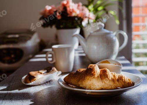 Preview: Breakfast at sunny morning. Croissant, tea and flower pot on the kitchen table. Light and shadows