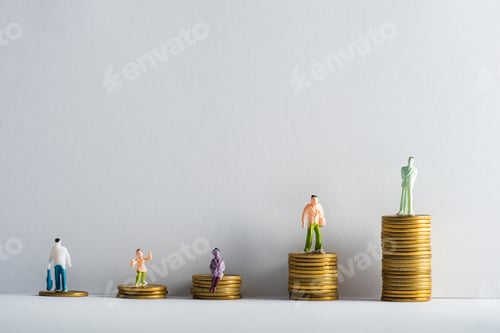 Preview: Plastic people figures on stacked golden coins on white surface on grey background, concept of