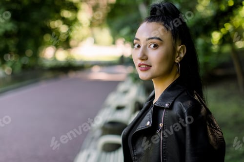 Preview: Attractive young Asian brunette woman in black leather jacket, relaxing on bench in the park outdoor