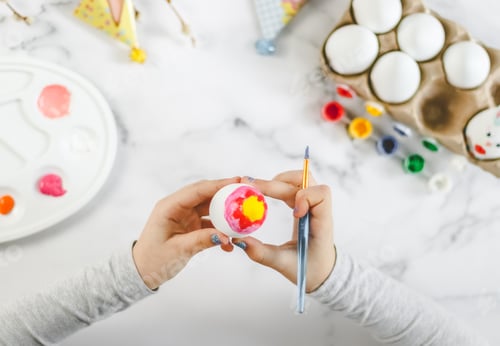 Preview: The hands of a girl hold an Easter egg with a brush with red and yellow acrylic paint