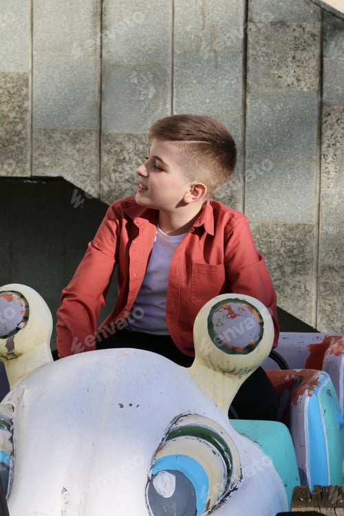 Preview: A blond boy with a fashionable haircut in a coral shirt and a white T-shirt is sitting in a toy car.