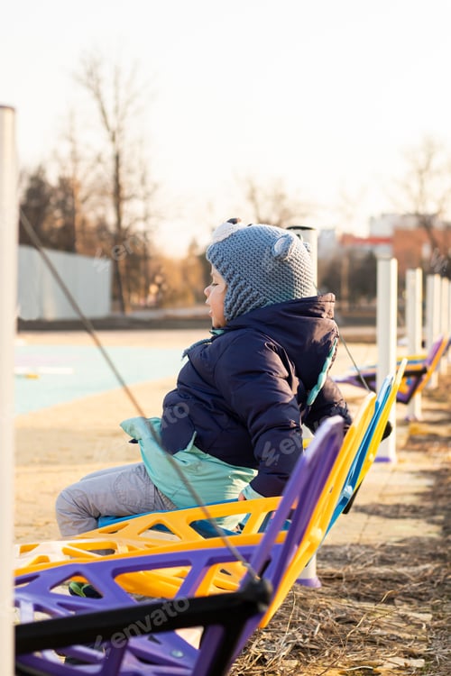 Preview: A little funny child rides on a colorful swing in a city park at sunset