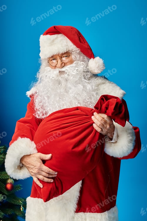 Preview: Santa Claus with beard and eyeglasses holding red sack bag with Christmas presents on blue